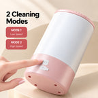 A hand presses the power button on a pink and white electric dog paw cleaner, showing its two cleaning modes and four holes on the base.