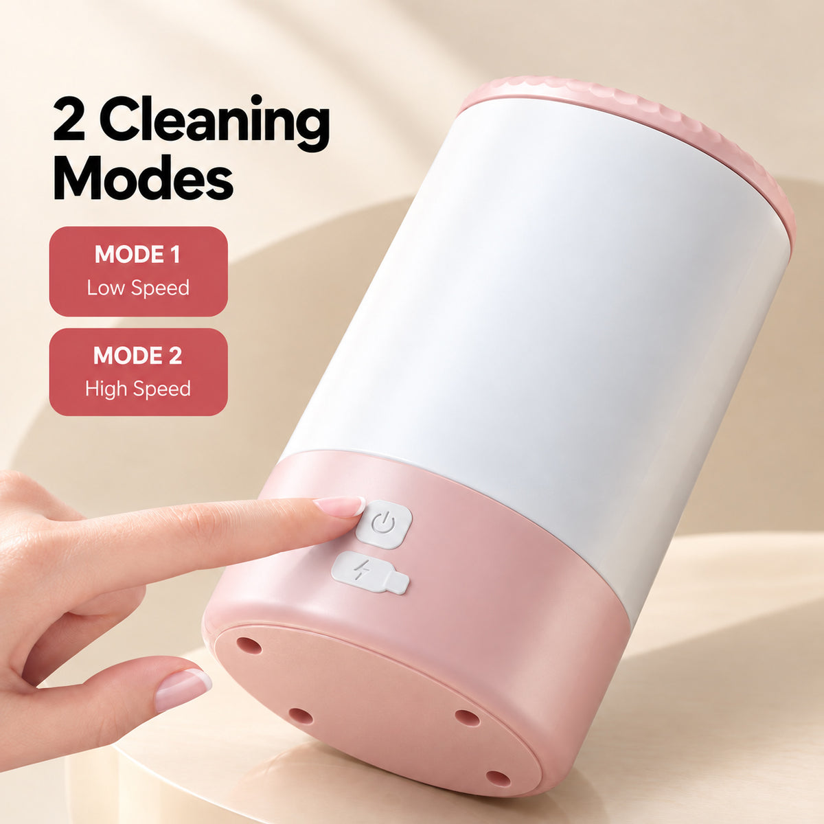 A hand presses the power button on a pink and white electric dog paw cleaner, showing its two cleaning modes and four holes on the base.